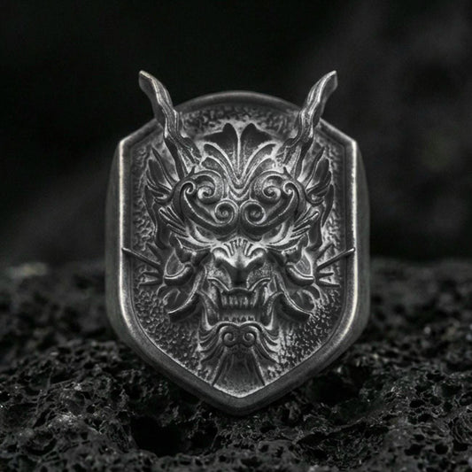 Oxidized sterling silver oni mask signet ring with horned Japanese demon face and deep relief texture, macro on black volcanic rock