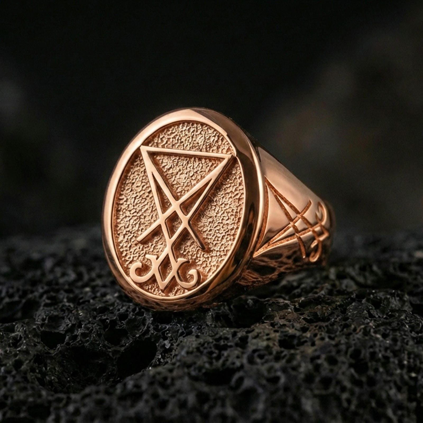 Rose gold plated Lucifer sigil signet ring with raised sigil emblem and matte-textured face, close-up on volcanic rock
