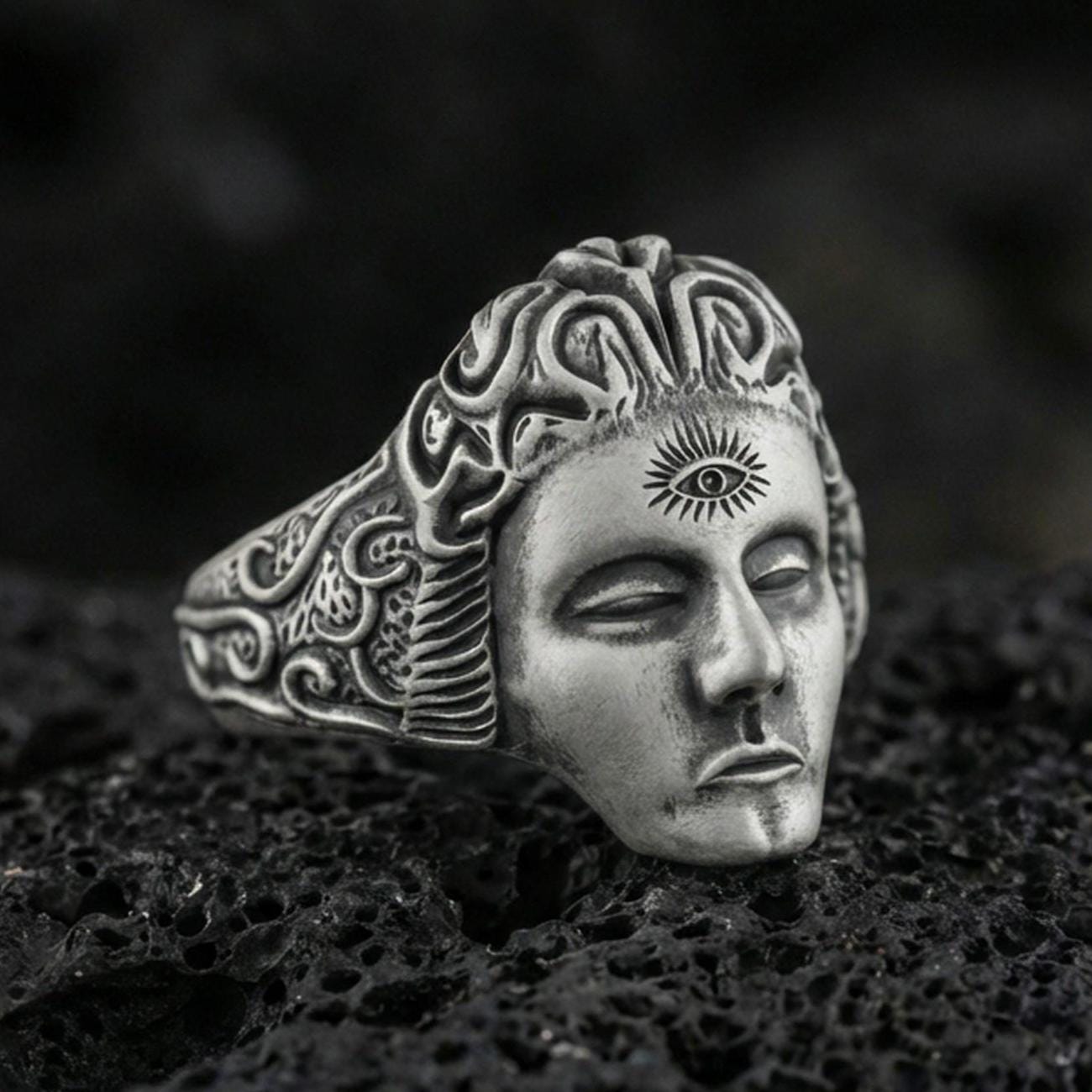 Sterling silver third eye face ring with an oxidized finish, sculpted calm mask and engraved swirling band details, macro on black volcanic rock