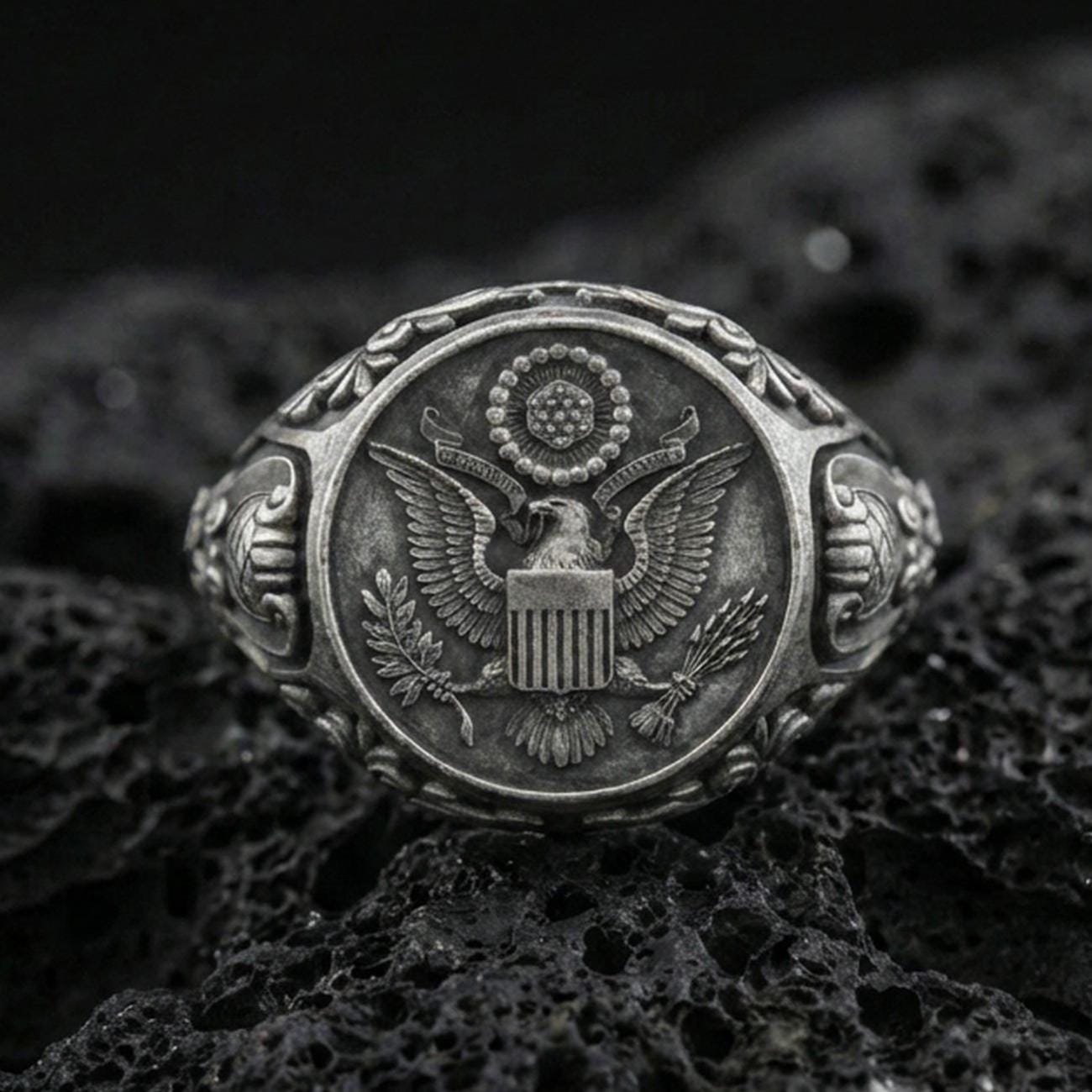 Sterling silver US eagle seal signet ring with an oxidized finish, detailed crest engraving and ornate shoulder accents, macro on black volcanic rock