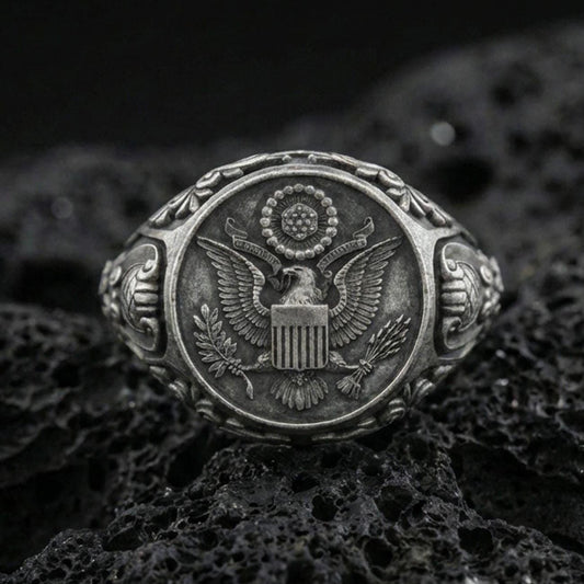 Sterling silver US eagle seal signet ring with an oxidized finish, detailed crest engraving and ornate shoulder accents, macro on black volcanic rock