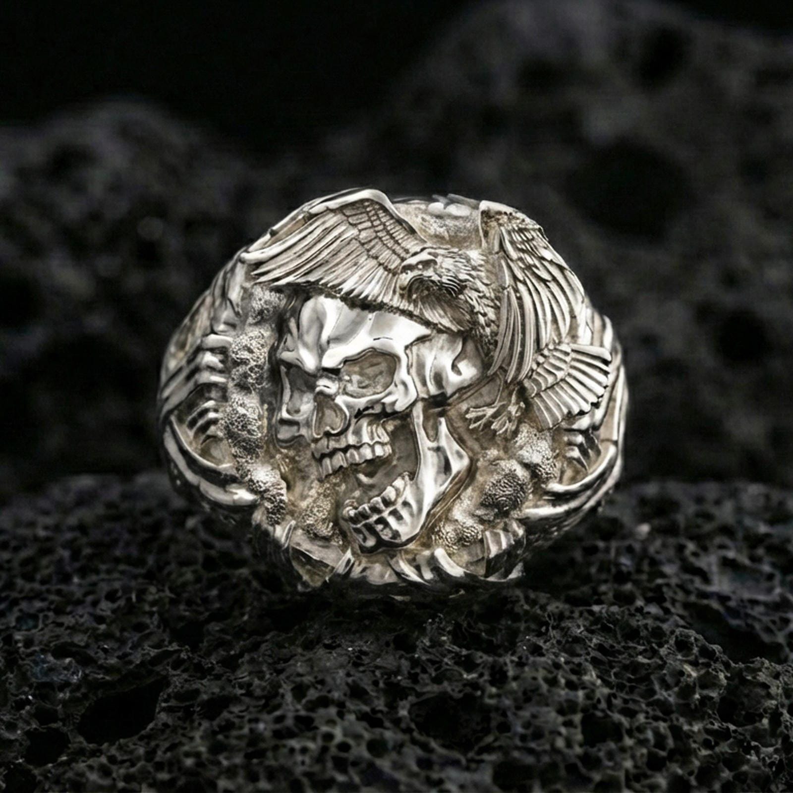 Sterling silver skull ring in a polished silver finish, high-relief eagle head and wing engraving with textured background, close-up on volcanic stone
