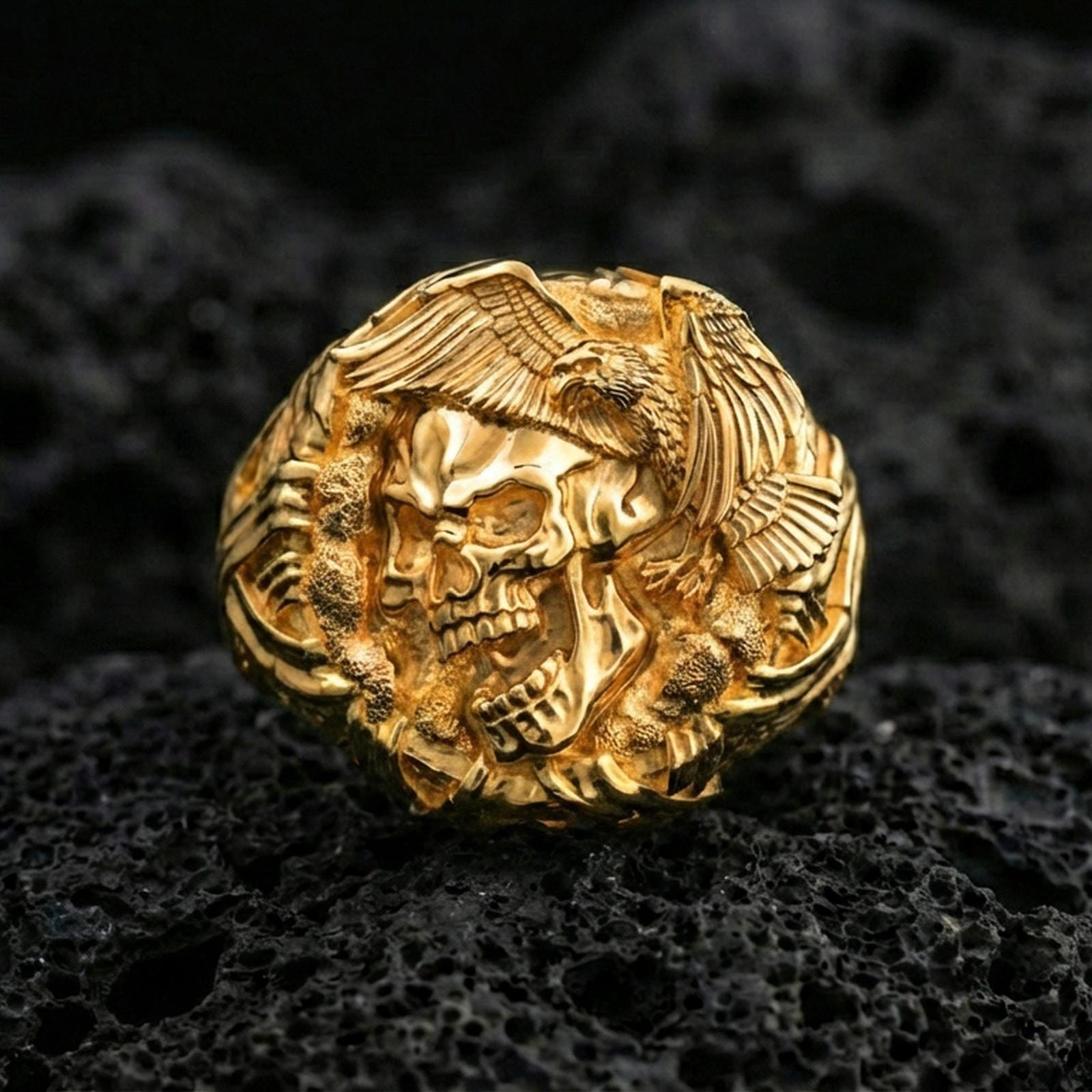Sterling silver skull ring with a gold plated finish, bold gothic skull centerpiece with eagle wings and talon accents, macro on volcanic rock