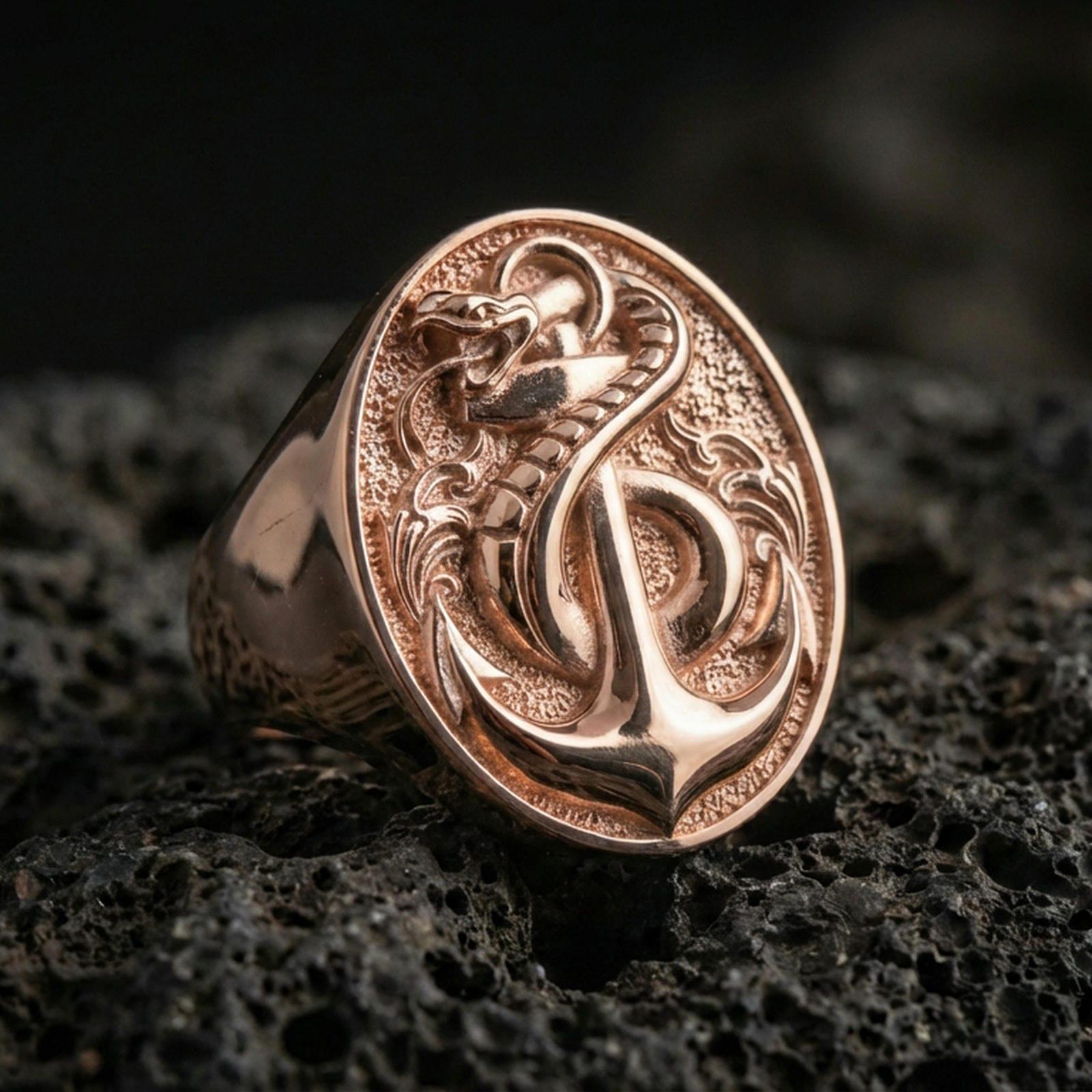 Sterling silver anchor and snake signet ring with a rose gold plated finish, raised anchor and coiled snake emblem, close-up on volcanic rock