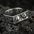 Load image into Gallery viewer, Sterling silver minimalist signet ring in a polished silver finish, rectangular recessed panel featuring skull motifs and relief texture, close-up on volcanic stone

