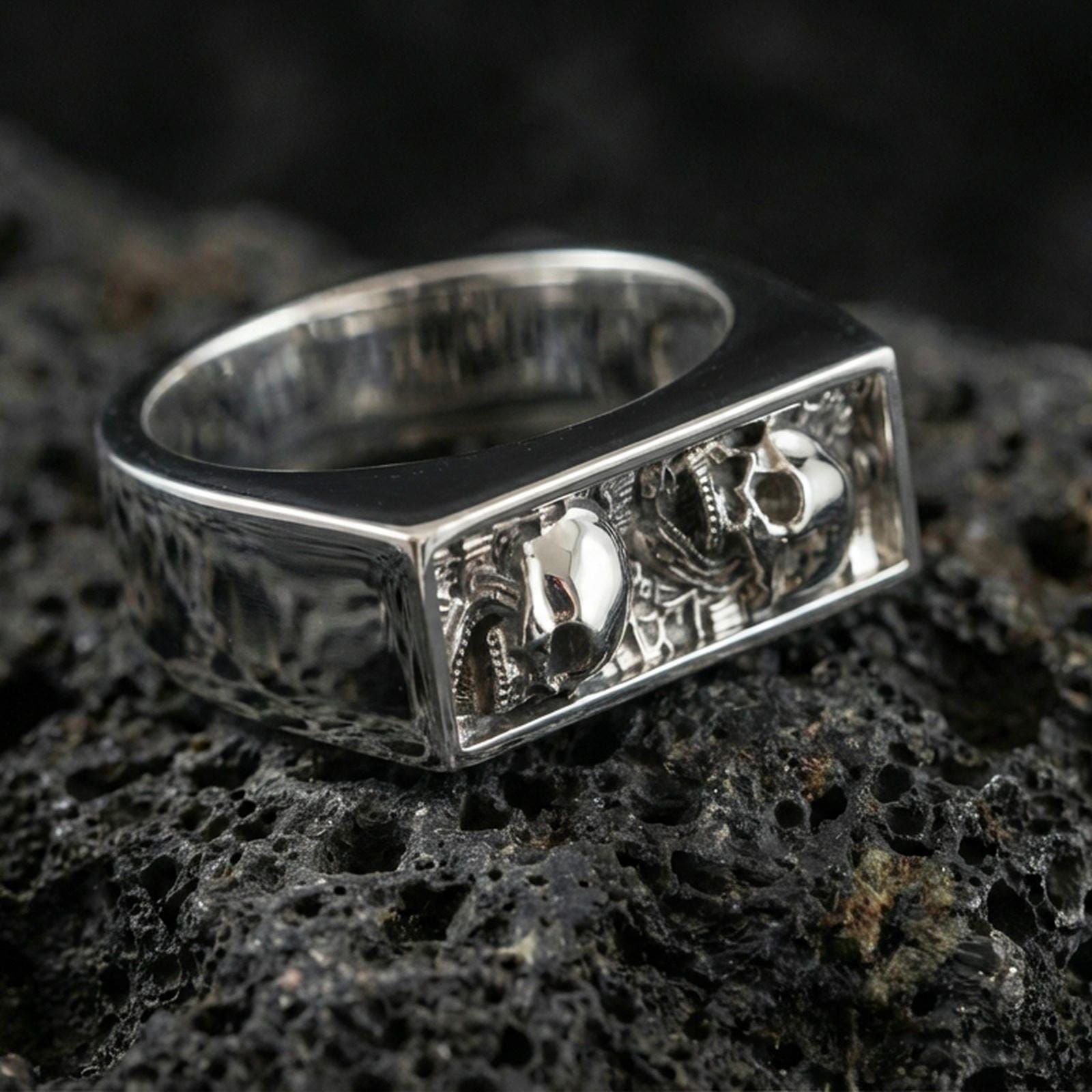 Sterling silver minimalist signet ring in a polished silver finish, rectangular recessed panel featuring skull motifs and relief texture, close-up on volcanic stone