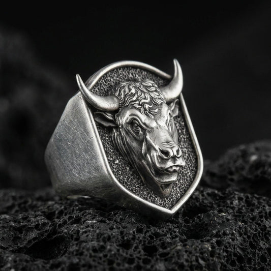 Oxidized sterling silver bull signet ring on textured volcanic rock, close-up macro showing horned head relief and shield-shaped face