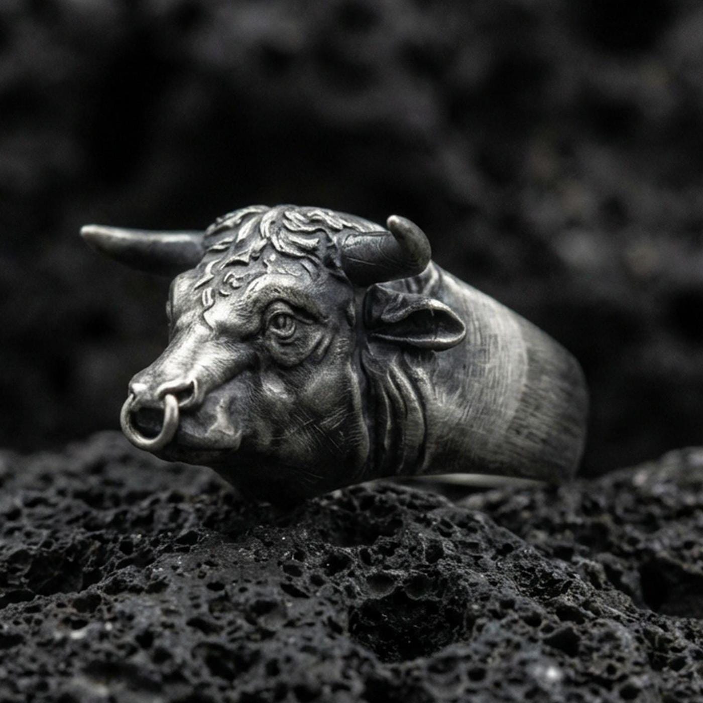 Oxidized sterling silver bull head ring on textured volcanic rock, macro close-up showing horns and nose ring detail in dark studio lighting