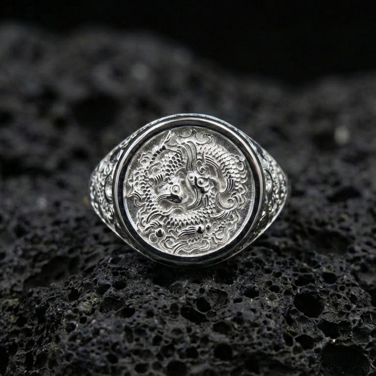 Polished silver koi fish ring on volcanic rock surface, close-up highlighting the round bezel and crisp engraved wave pattern