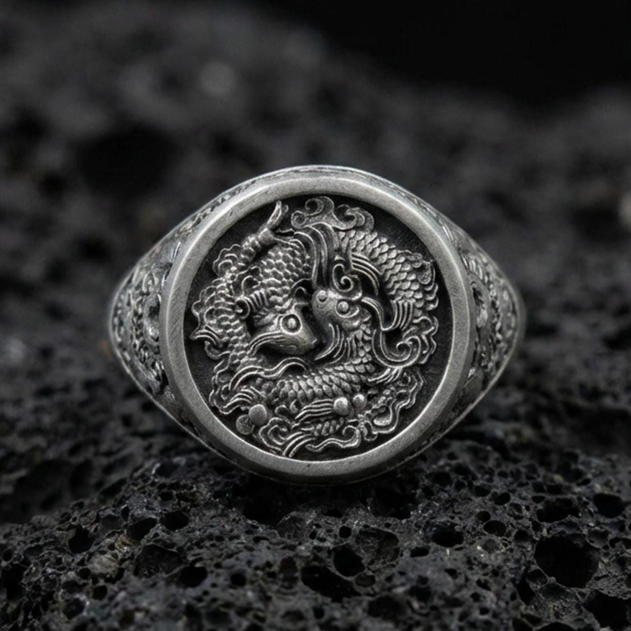 Oxidized sterling silver koi fish signet ring on textured volcanic rock, macro close-up showing detailed Japanese-inspired scales and swirling water relief