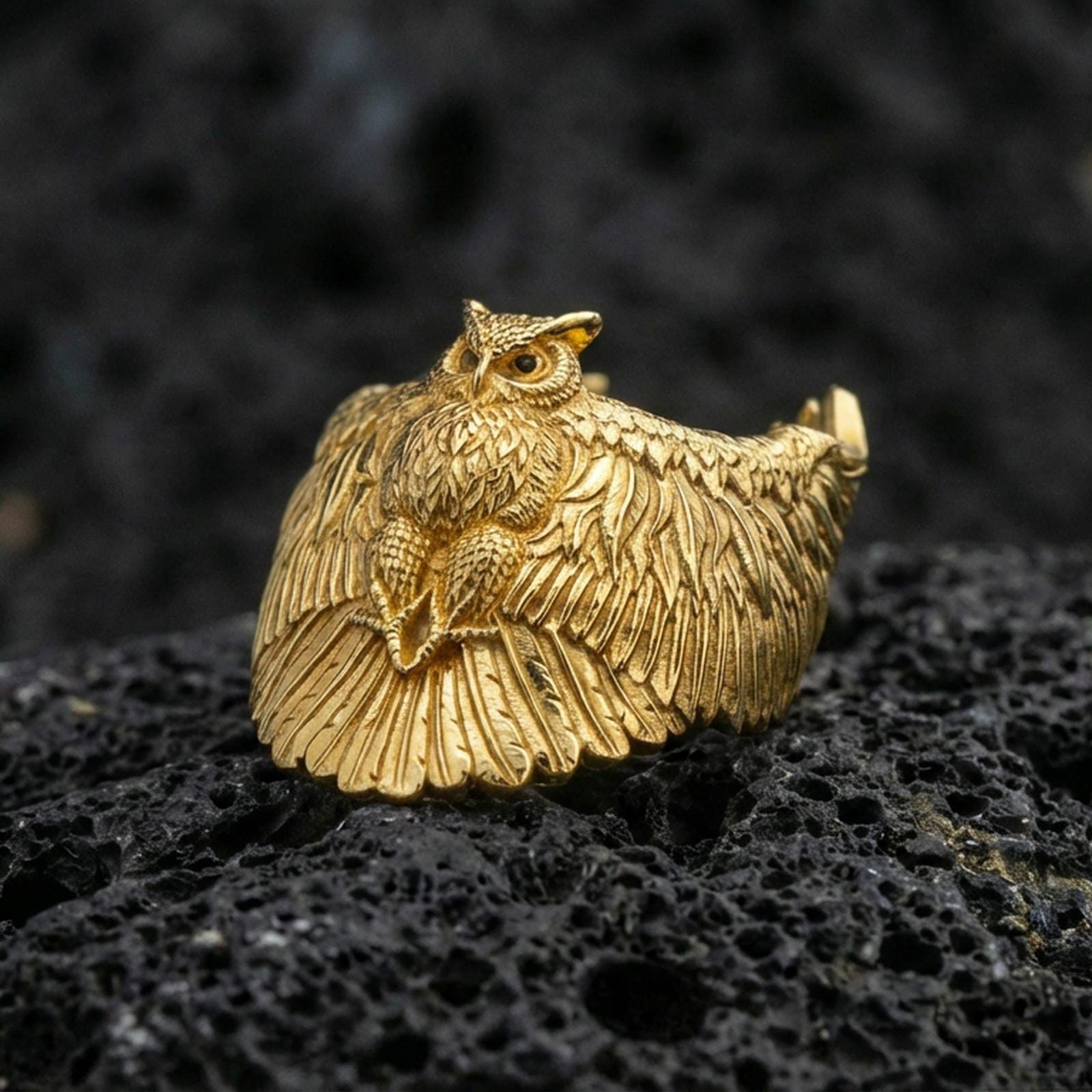 Gold plated owl ring on porous volcanic stone, premium dark studio shot emphasizing the owl body relief and layered wing feathers