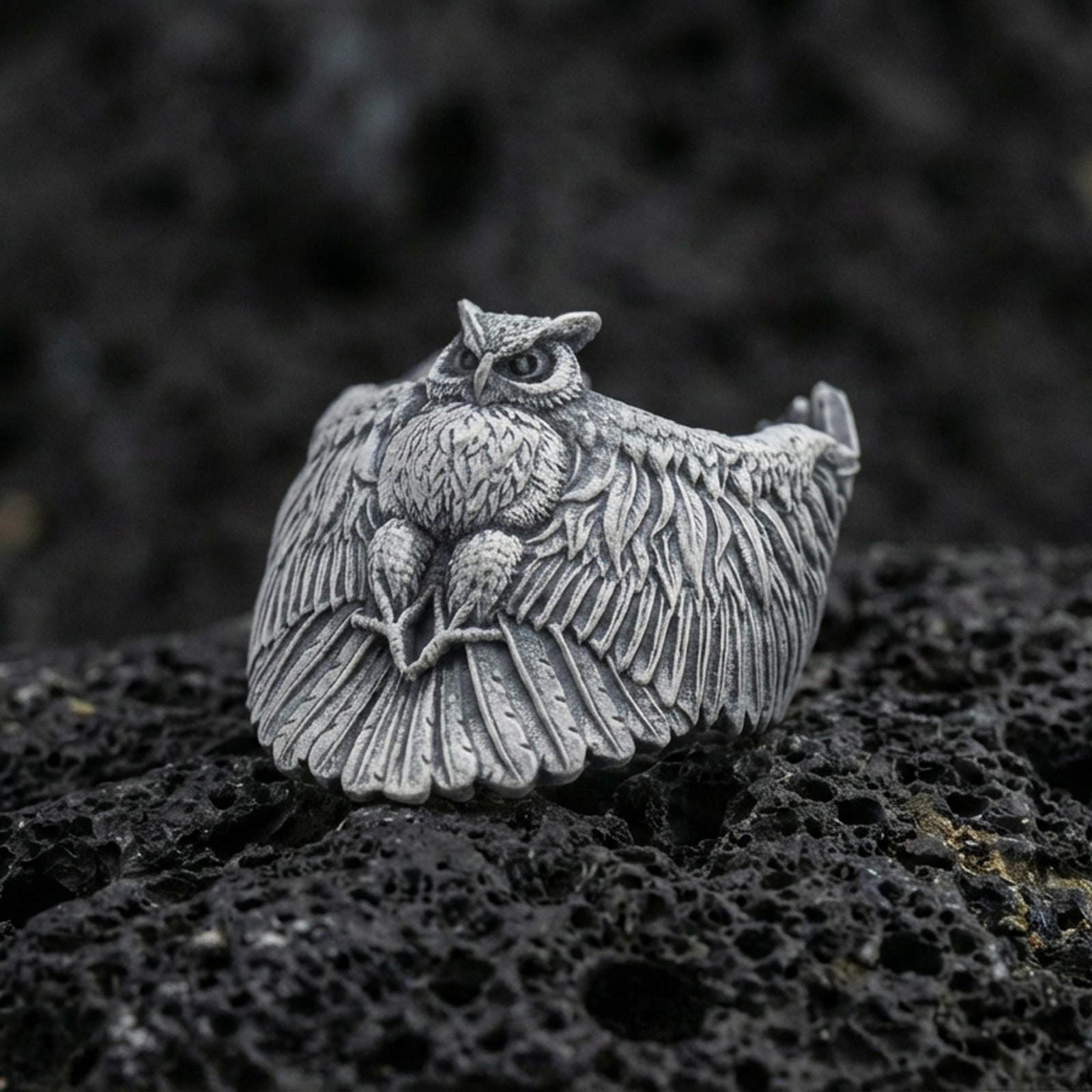 Oxidized sterling silver owl ring on textured volcanic rock, macro close-up showing spread wings, feather engraving, and intense owl eyes