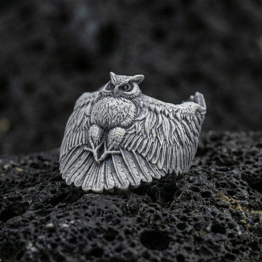 Oxidized sterling silver owl ring on textured volcanic rock, macro close-up showing spread wings, feather engraving, and intense owl eyes