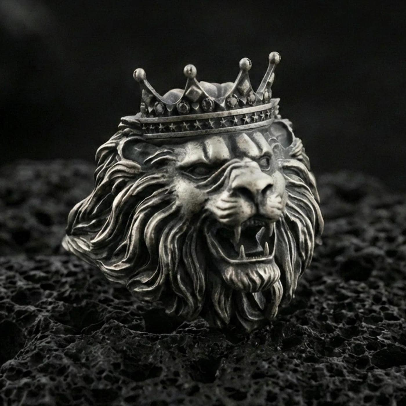 Oxidized sterling silver crowned lion ring on textured volcanic rock, macro close-up showing roaring lion head, detailed mane, and royal crown