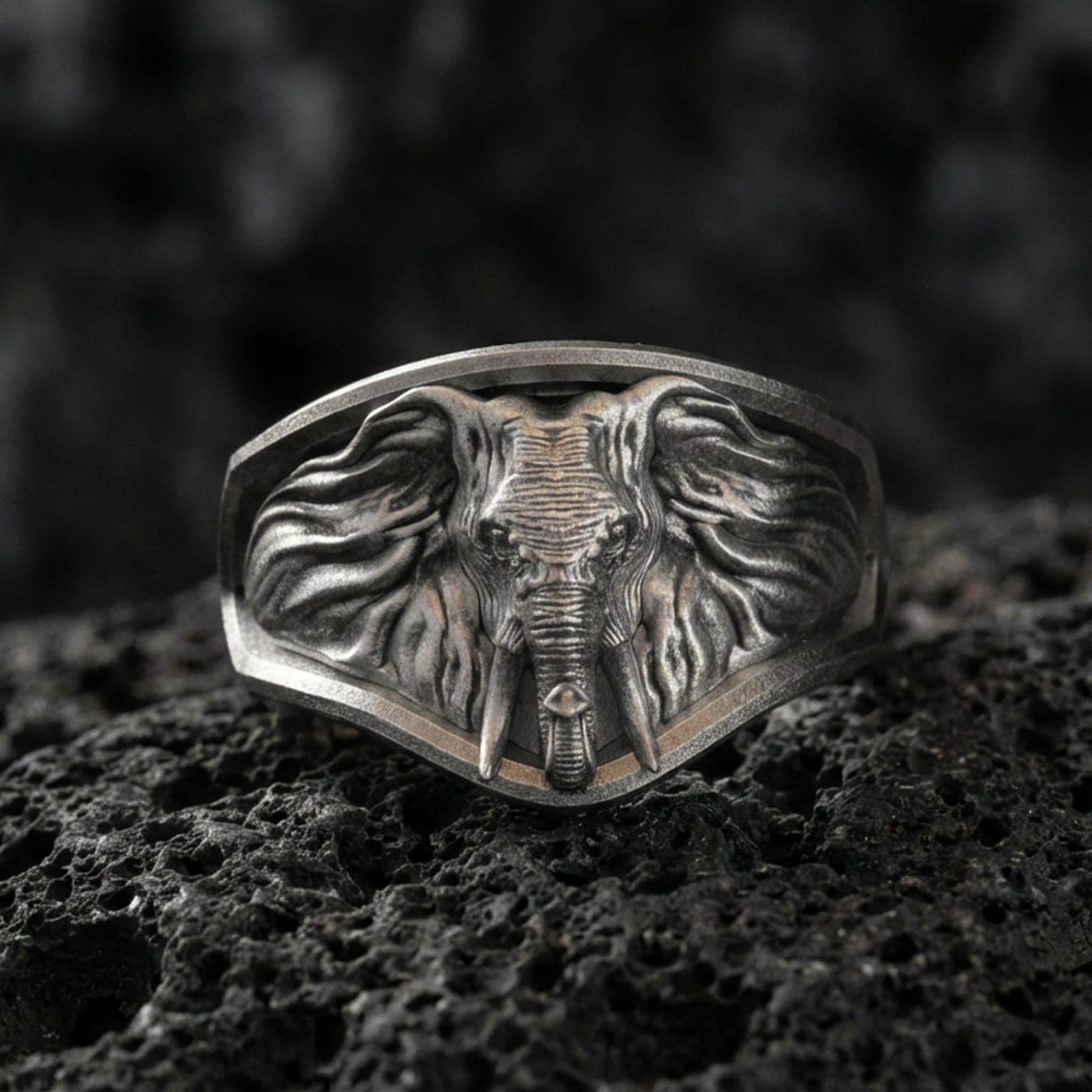 Oxidized sterling silver elephant signet ring on textured volcanic rock, macro close-up showing frontal elephant face, tusks, and flowing ear details