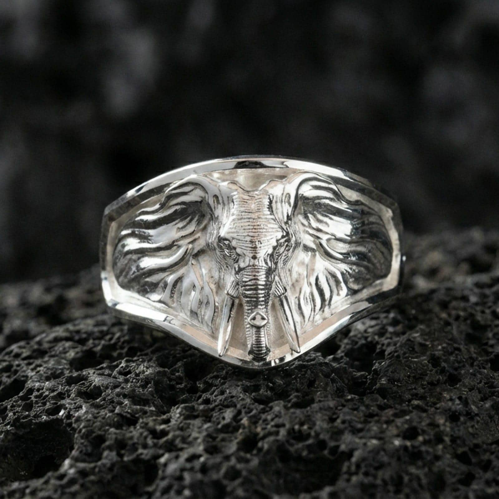 Polished silver elephant head signet ring on volcanic rock surface, close-up highlighting the wide geometric face, sculpted trunk, and crisp bezel edges