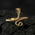 Load image into Gallery viewer, Gold plated cobra snake ring on porous volcanic stone, premium dark studio shot emphasizing the upright hood, curled tail, and realistic texture
