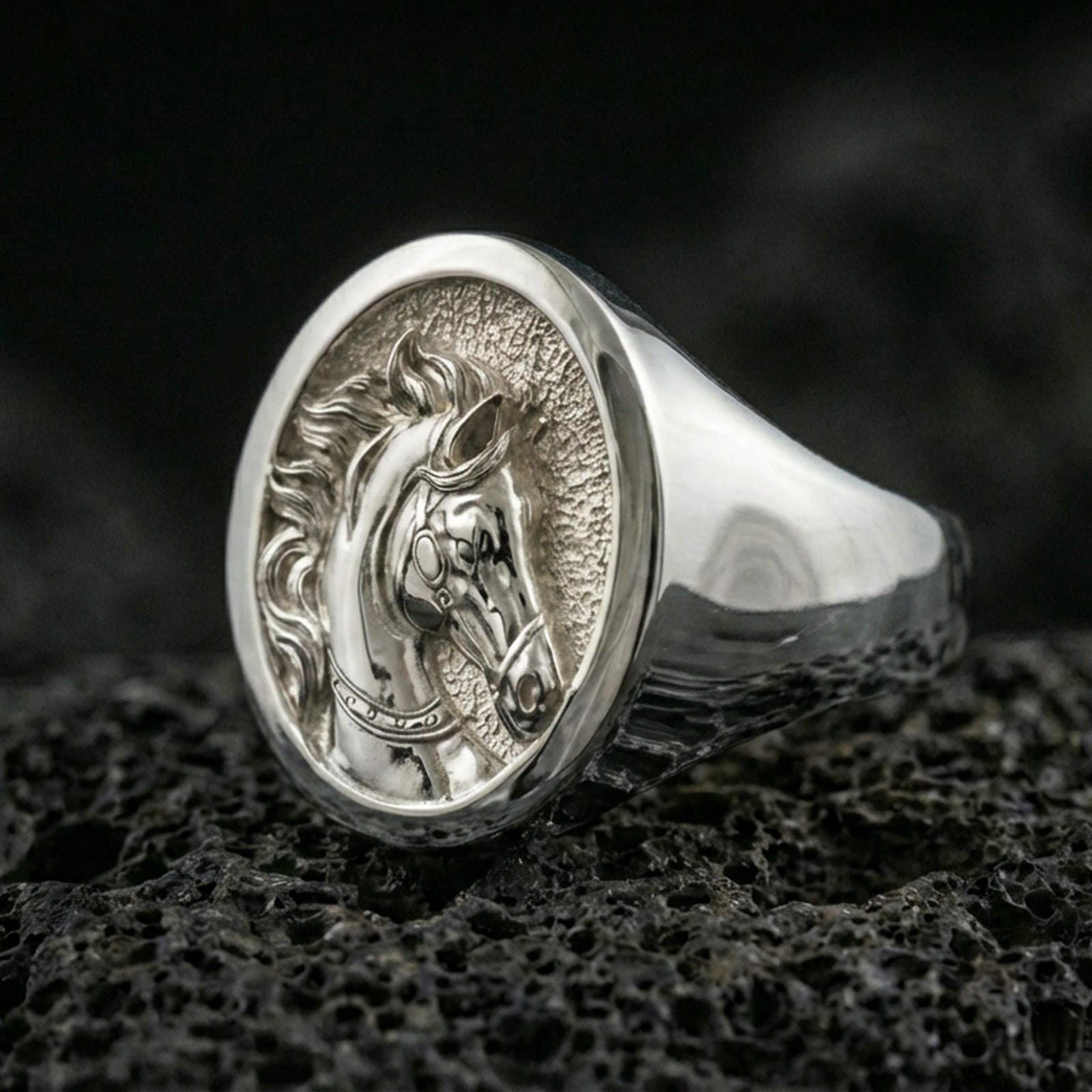 Polished silver horse head signet ring on volcanic rock surface, close-up highlighting the round bezel, carved mane detail, and smooth tapered band