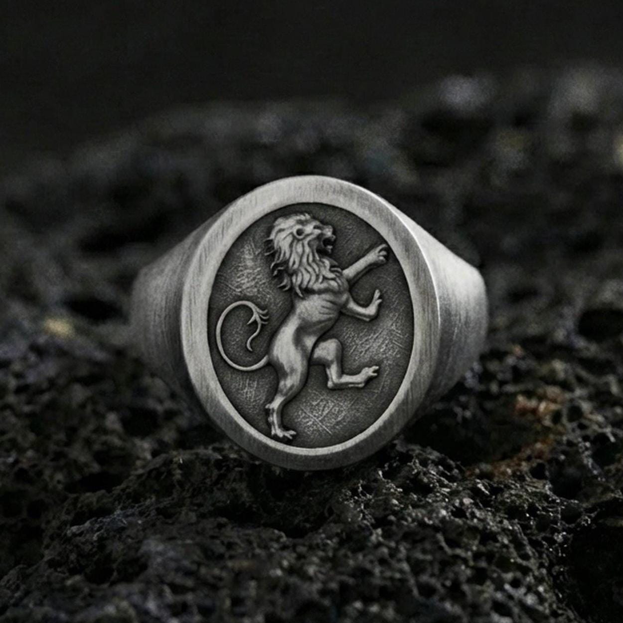 Oxidized sterling silver heraldic lion signet ring on black volcanic rock, macro close-up highlighting the crowned lion emblem and dark recessed background