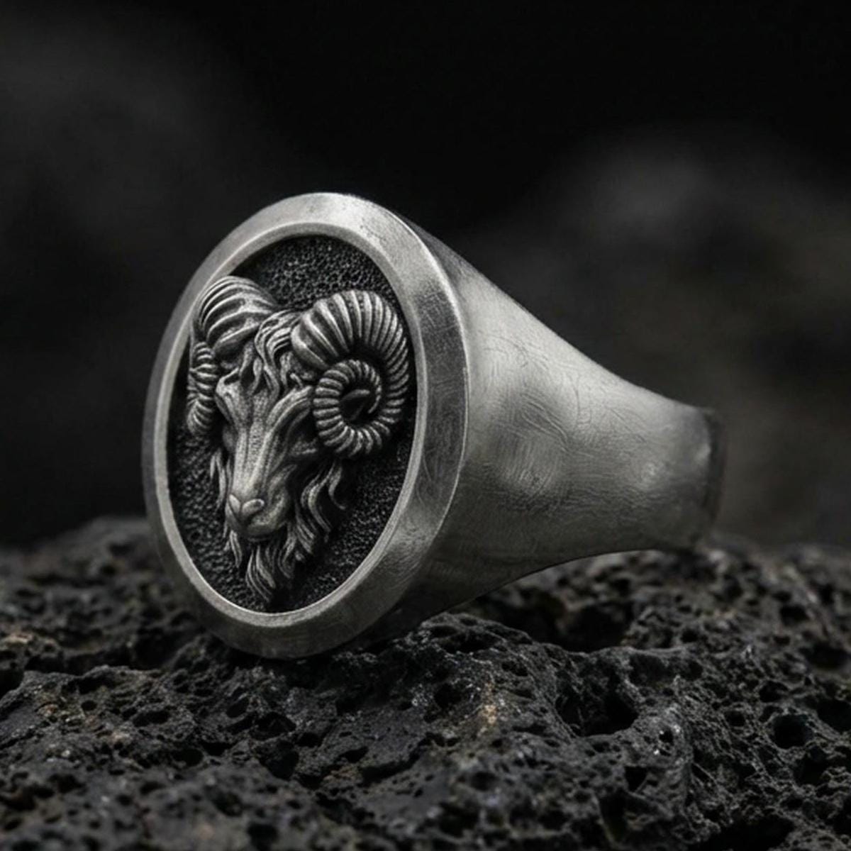 Close-up of an oxidized sterling silver ram signet ring on porous volcanic rock with dark premium lighting