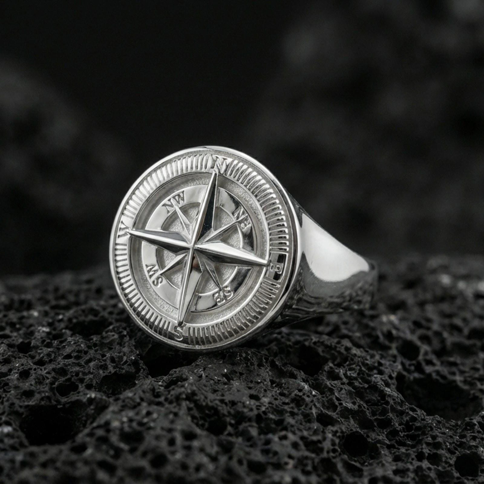 Sterling silver compass ring in a polished silver finish, round signet face with detailed compass rose and directional markings, close-up on volcanic stone