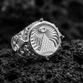 Load image into Gallery viewer, Sterling silver illuminati-inspired signet ring in a polished silver finish, round face with eye above pyramid and swirling engraved background, close-up on volcanic stone
