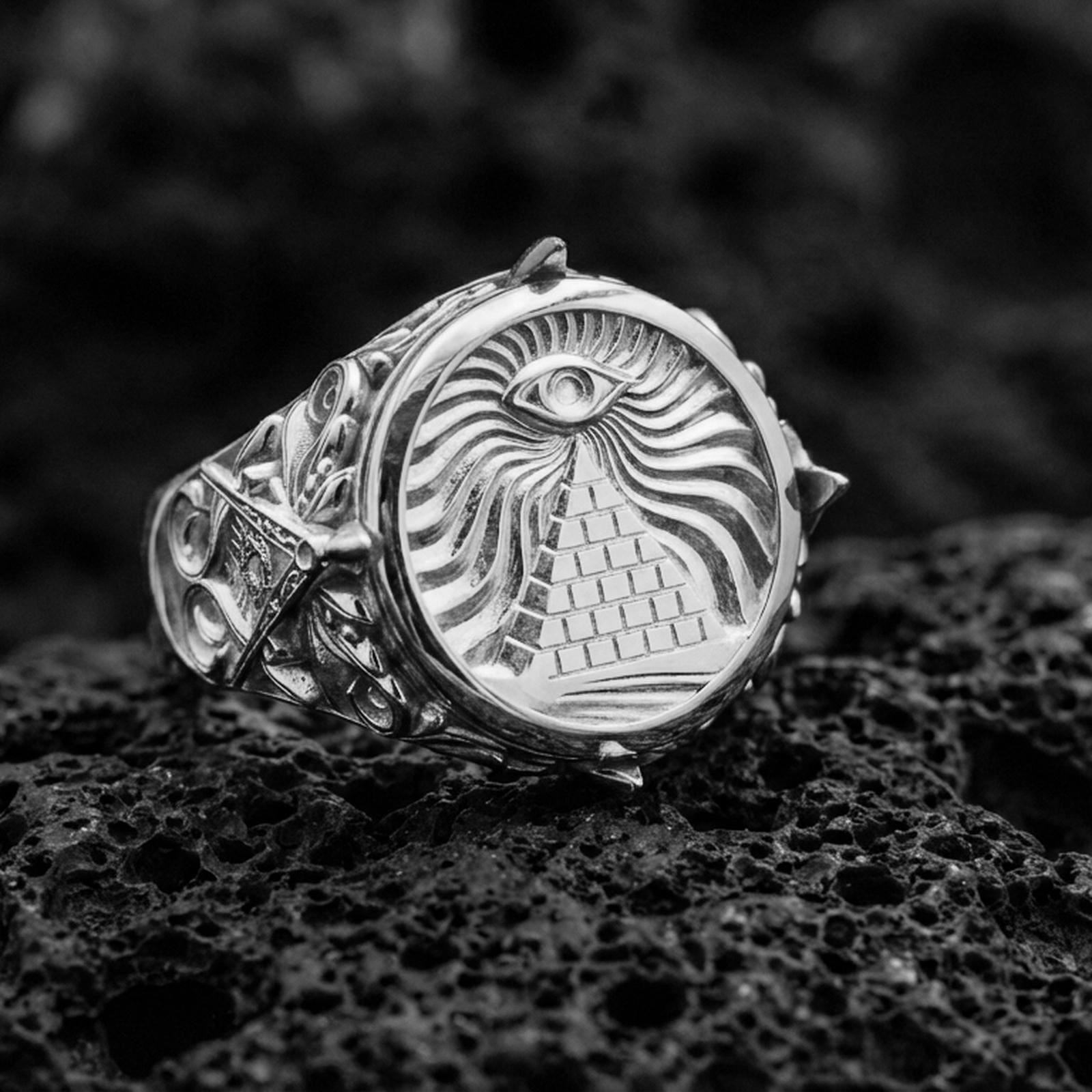 Sterling silver illuminati-inspired signet ring in a polished silver finish, round face with eye above pyramid and swirling engraved background, close-up on volcanic stone