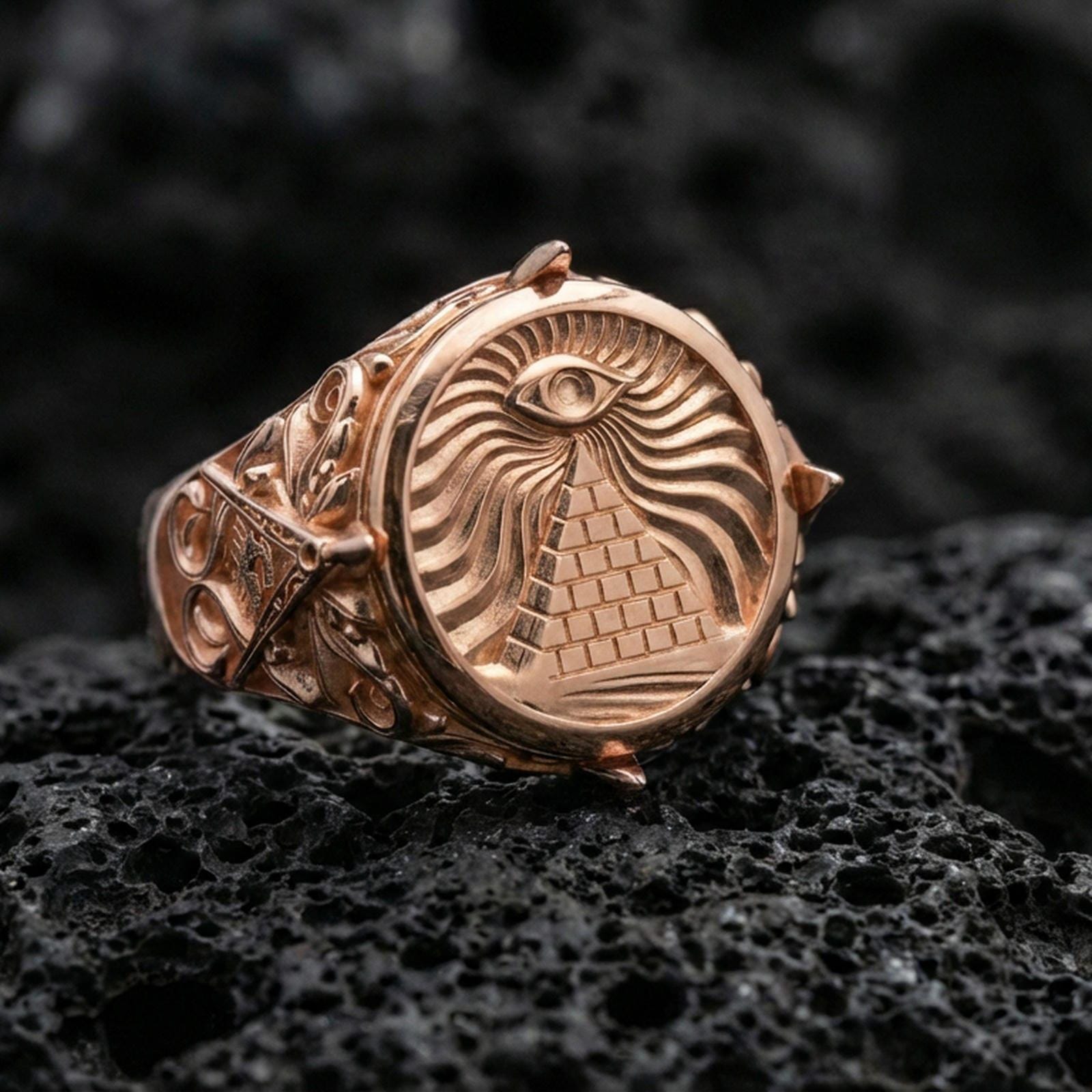 Sterling silver pyramid eye signet ring with a rose gold plated finish, bold round medallion face and engraved mystical rays, close-up on volcanic rock