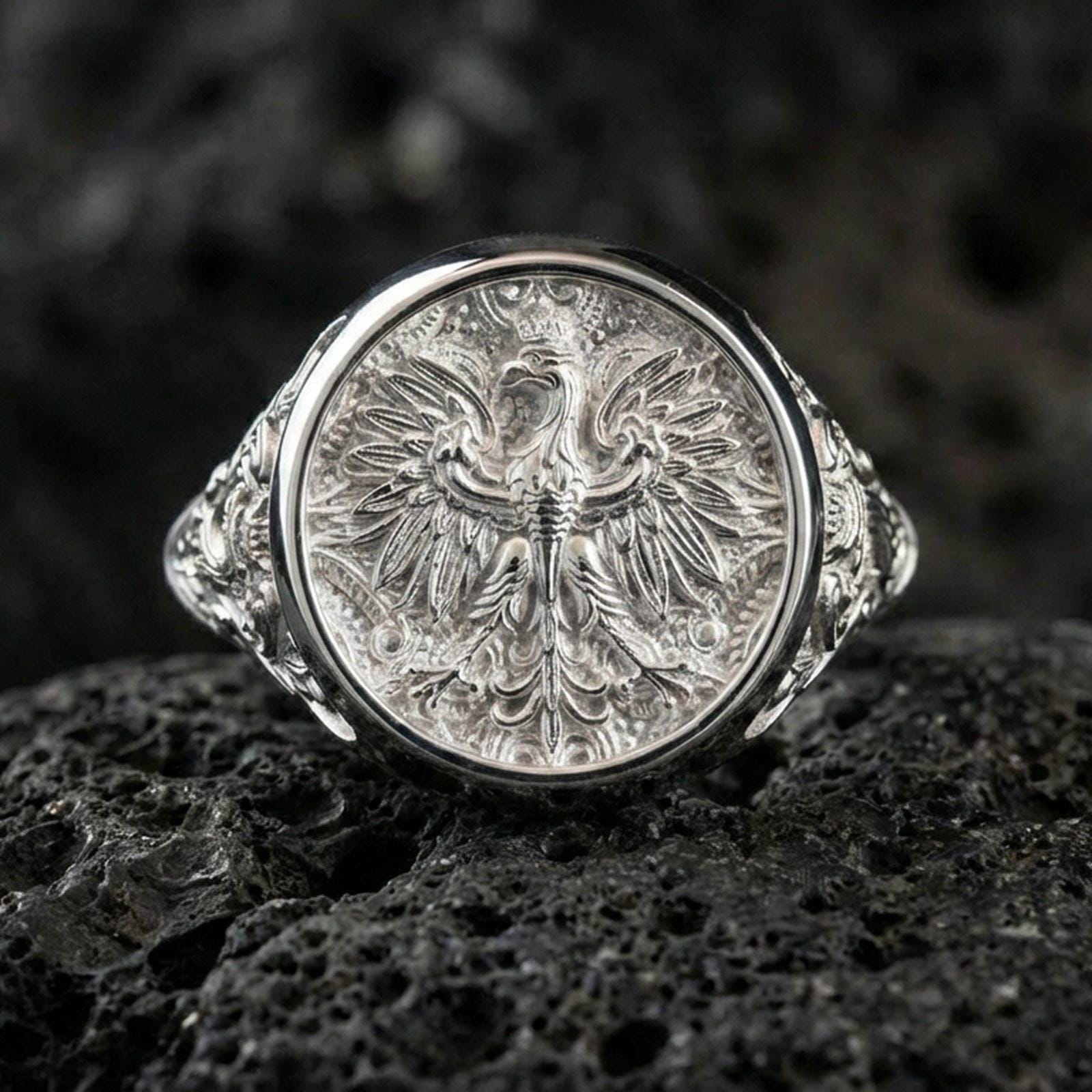 Sterling silver phoenix crest ring in a polished silver finish, ornate round signet design with layered feather engraving, close-up on volcanic stone