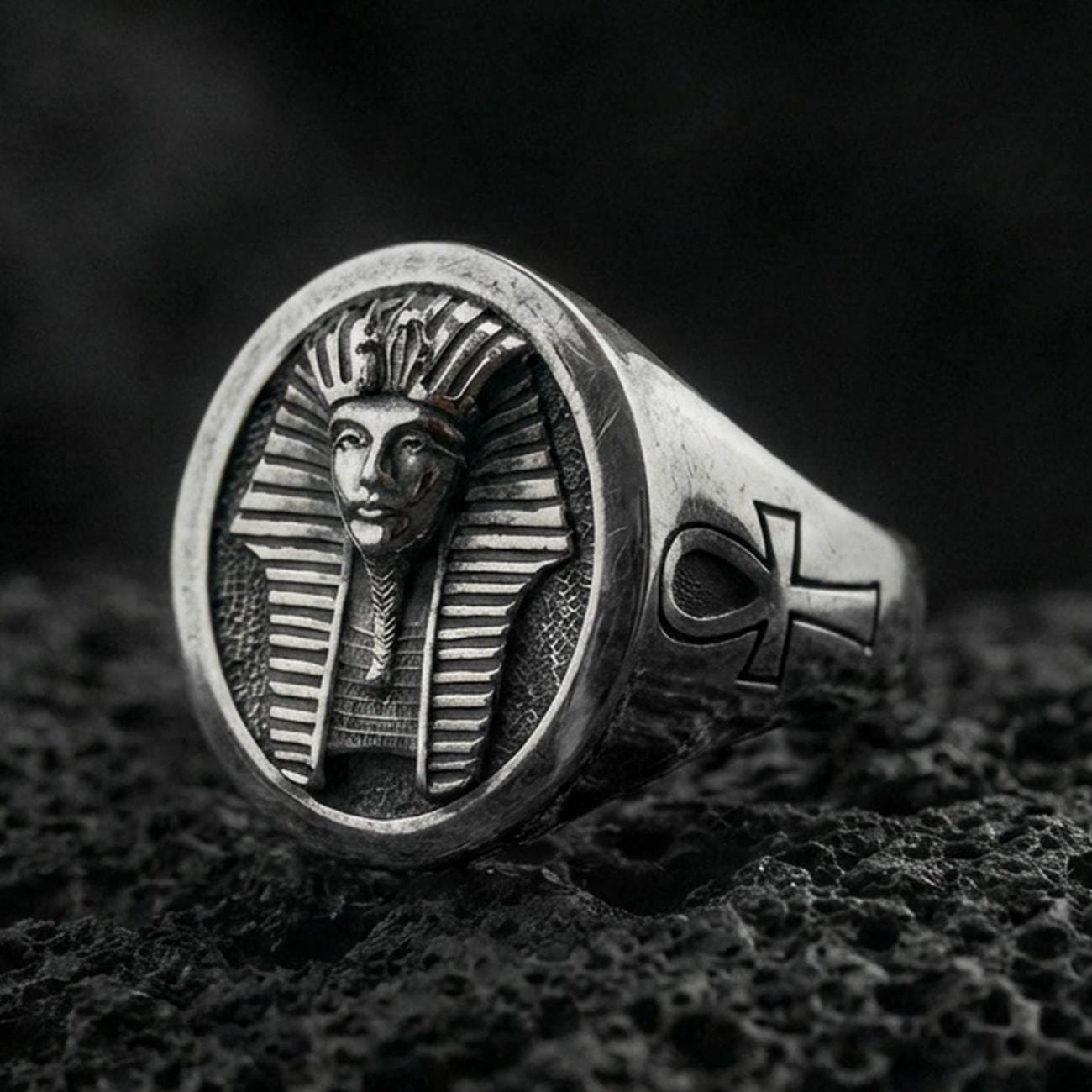 Sterling silver Egyptian pharaoh signet ring with an oxidized finish, oval face featuring a carved ancient king bust and side ankh symbol, macro on black volcanic rock