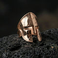Load image into Gallery viewer, Sterling silver gladiator-style helmet ring with a rose gold plated finish, sculpted Spartan warrior helmet face and distressed crack detailing, close-up on black volcanic rock
