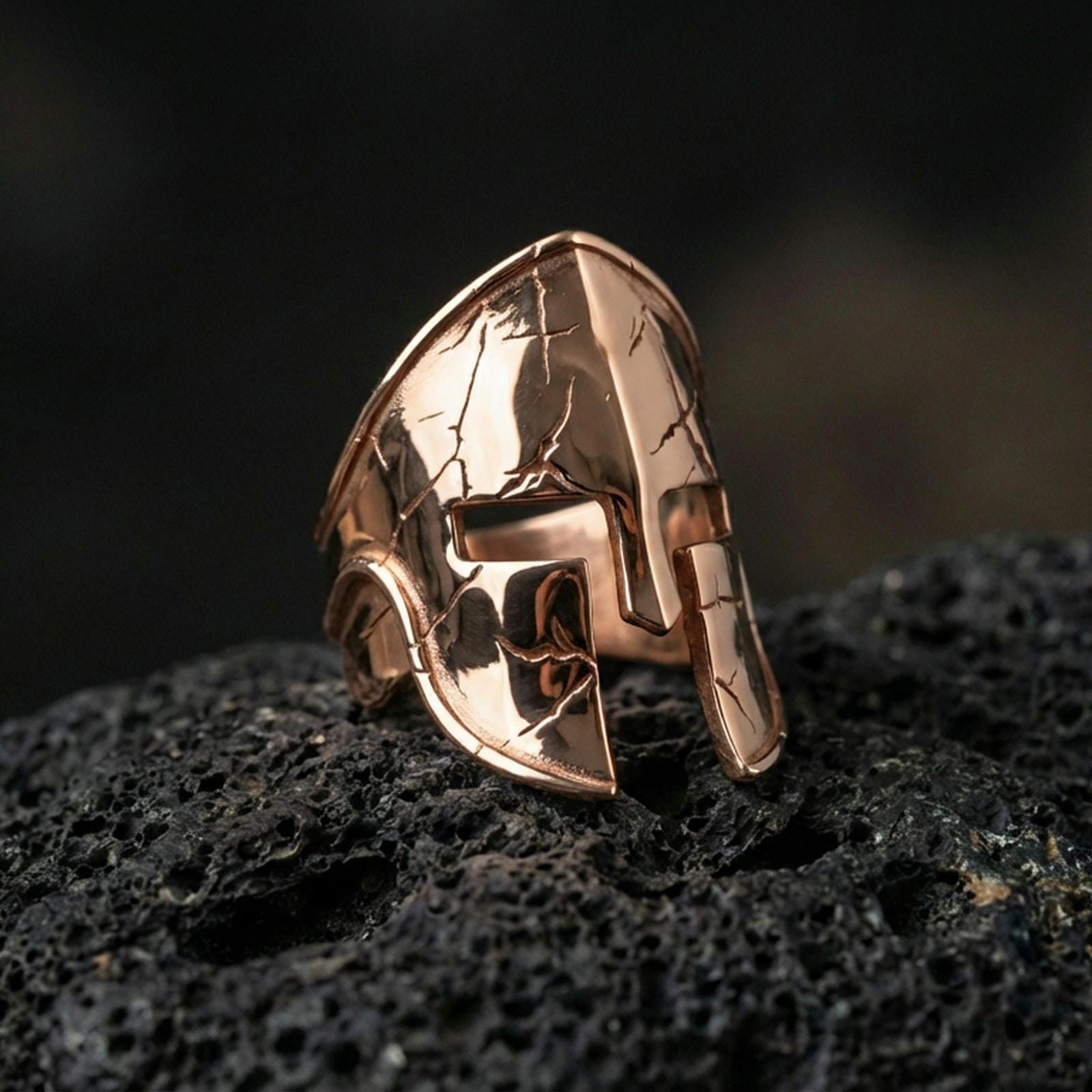 Sterling silver gladiator-style helmet ring with a rose gold plated finish, sculpted Spartan warrior helmet face and distressed crack detailing, close-up on black volcanic rock