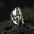 Load image into Gallery viewer, Sterling silver Spartan helmet ring in a polished silver finish, bold sculptural warrior helm face with engraved crack patterns, close-up on volcanic stone
