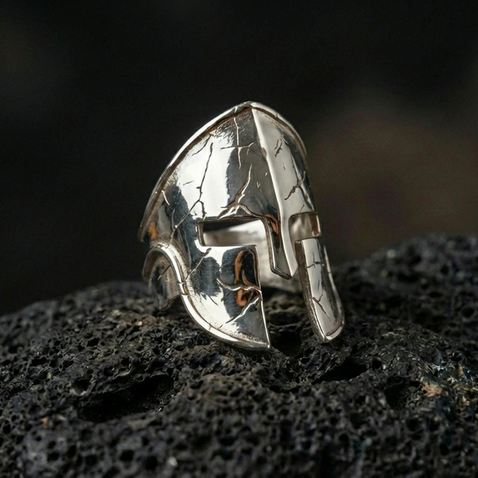 Sterling silver Spartan helmet ring in a polished silver finish, bold sculptural warrior helm face with engraved crack patterns, close-up on volcanic stone