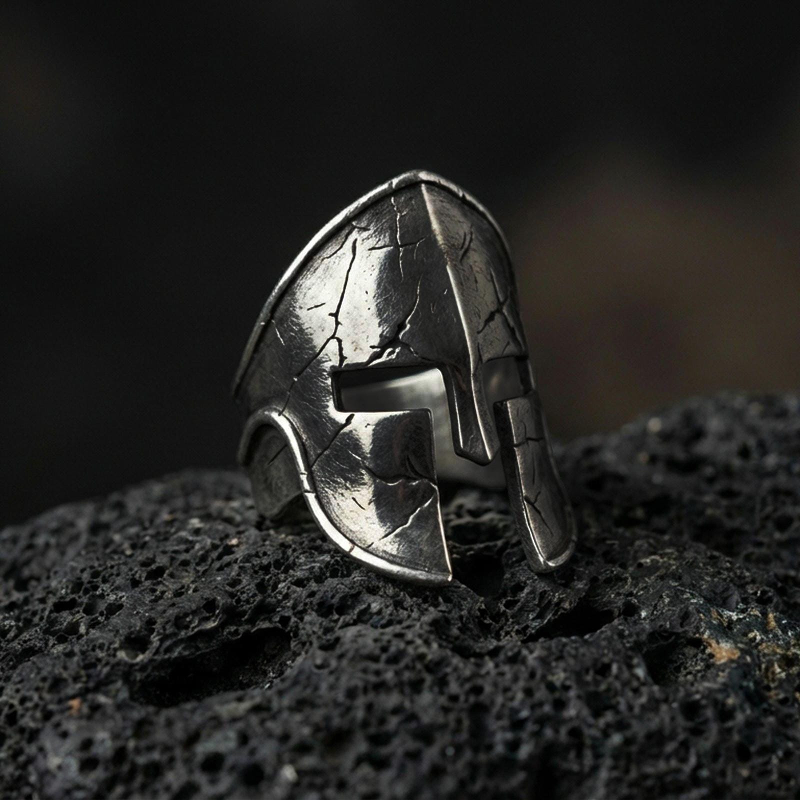 Sterling silver Spartan warrior helmet signet ring with an oxidized finish, ancient Greek helmet design with cracked texture detail, macro on black volcanic rock