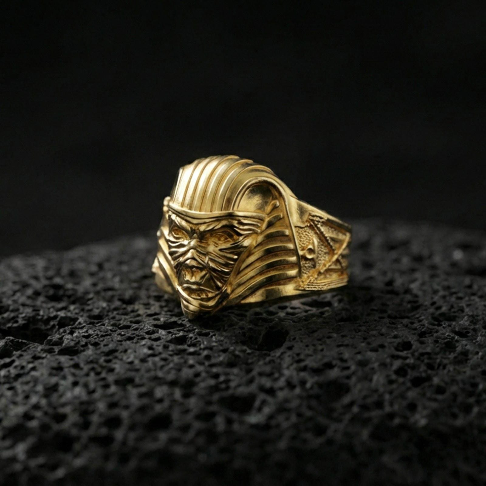 Sterling silver pharaoh mummy ring with a gold plated finish, detailed undead mask design and engraved side symbols, macro product shot on volcanic rock