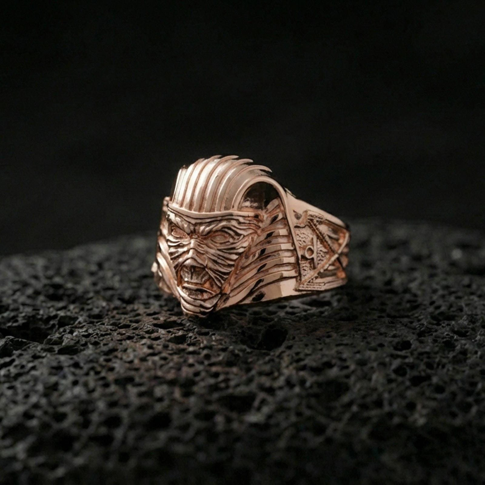 Sterling silver ancient Egypt horror mask ring with a rose gold plated finish, bold mummy pharaoh face and textured headdress lines, close-up on black volcanic rock