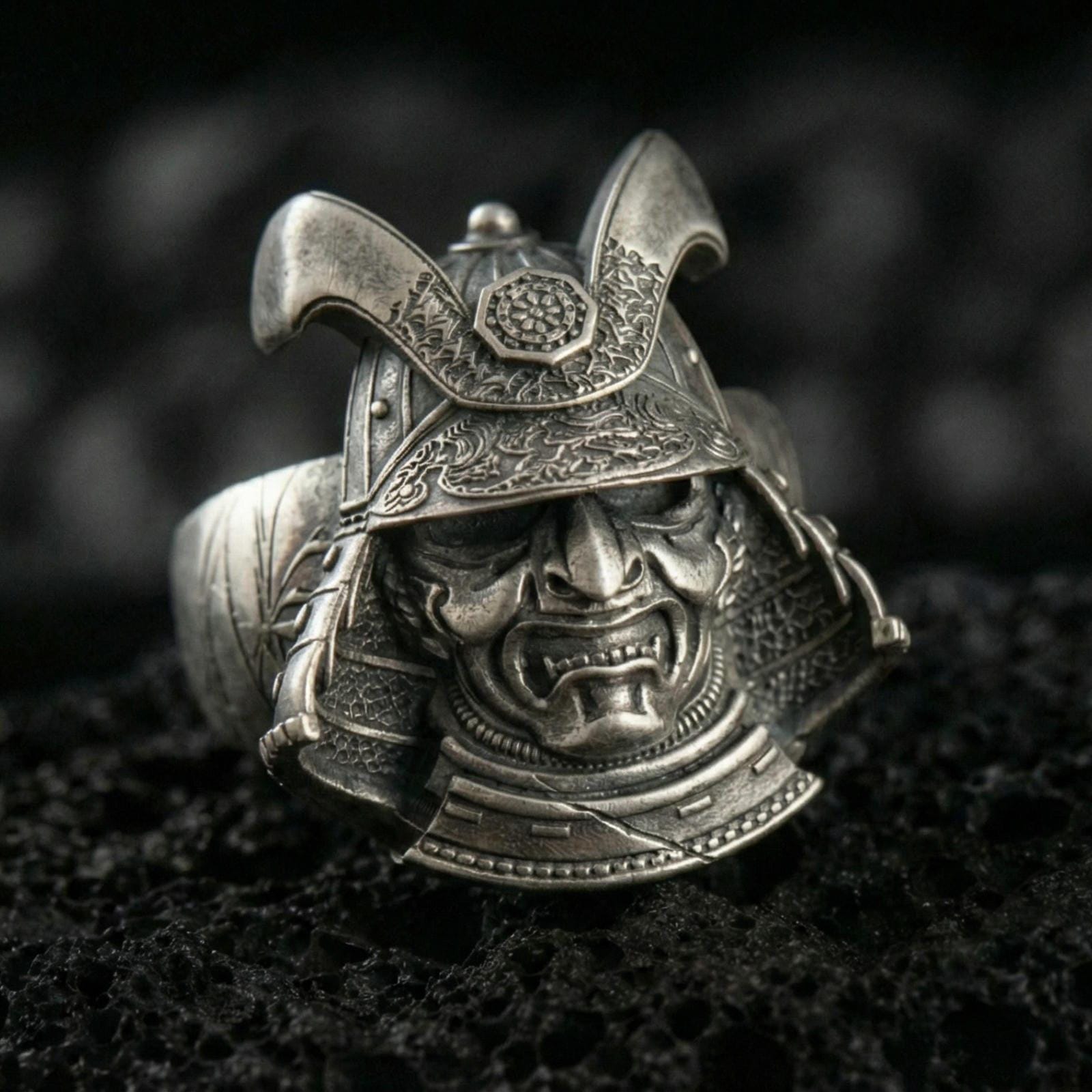 Sterling silver samurai mask ring with an oxidized finish, fierce warrior face and engraved kabuto helmet details, macro on black volcanic rock