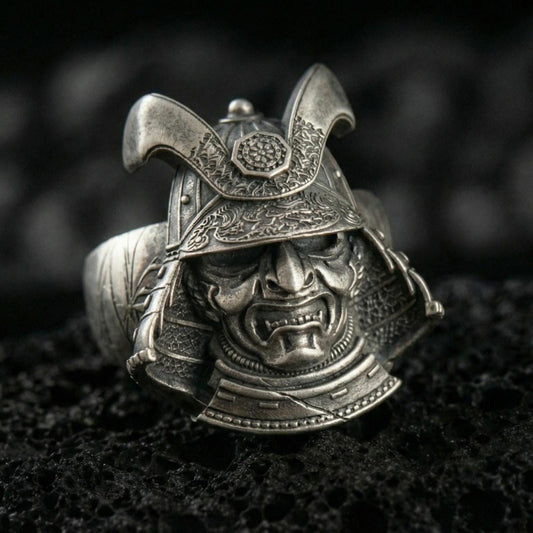 Sterling silver samurai mask ring with an oxidized finish, fierce warrior face and engraved kabuto helmet details, macro on black volcanic rock