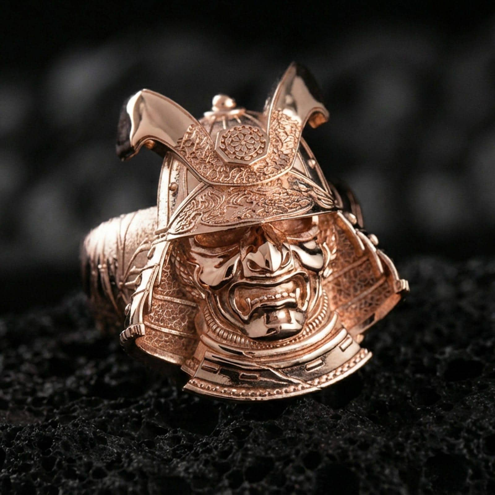 Sterling silver samurai mask statement ring with a rose gold plated finish, bold Japanese warrior helmet motif and engraved ornamental detailing, close-up on black volcanic rock