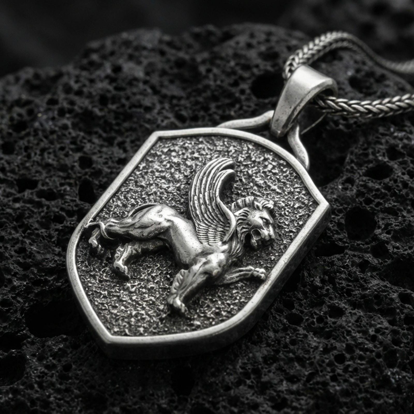 Sterling silver winged lion shield pendant with an oxidized finish, medieval heraldic griffin-style motif in raised relief, close-up on black volcanic rock