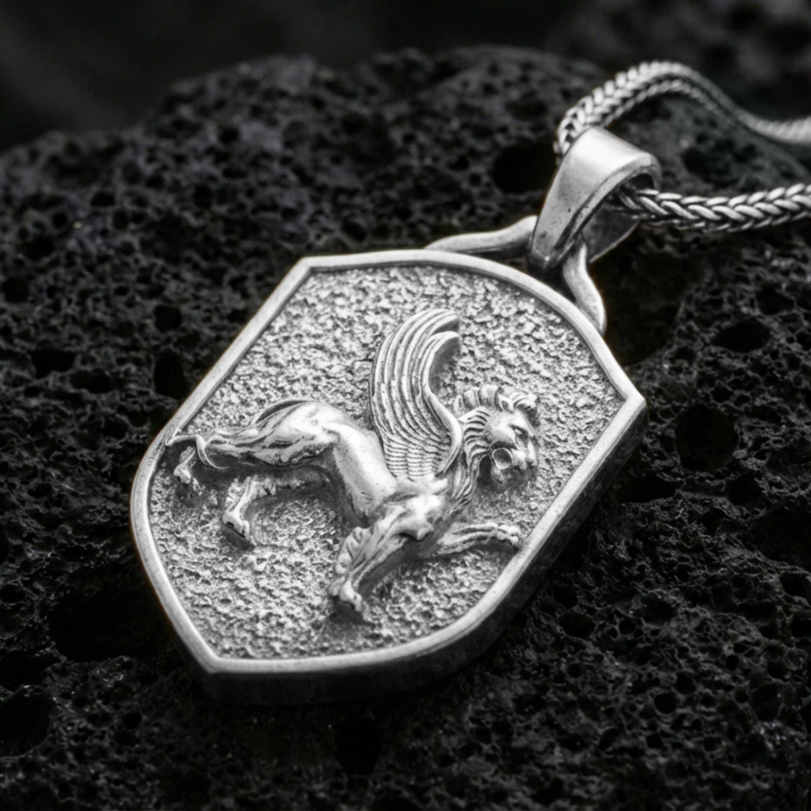 Sterling silver winged lion crest pendant in a polished silver finish, shield-shaped heraldic animal emblem with textured background, macro on volcanic stone