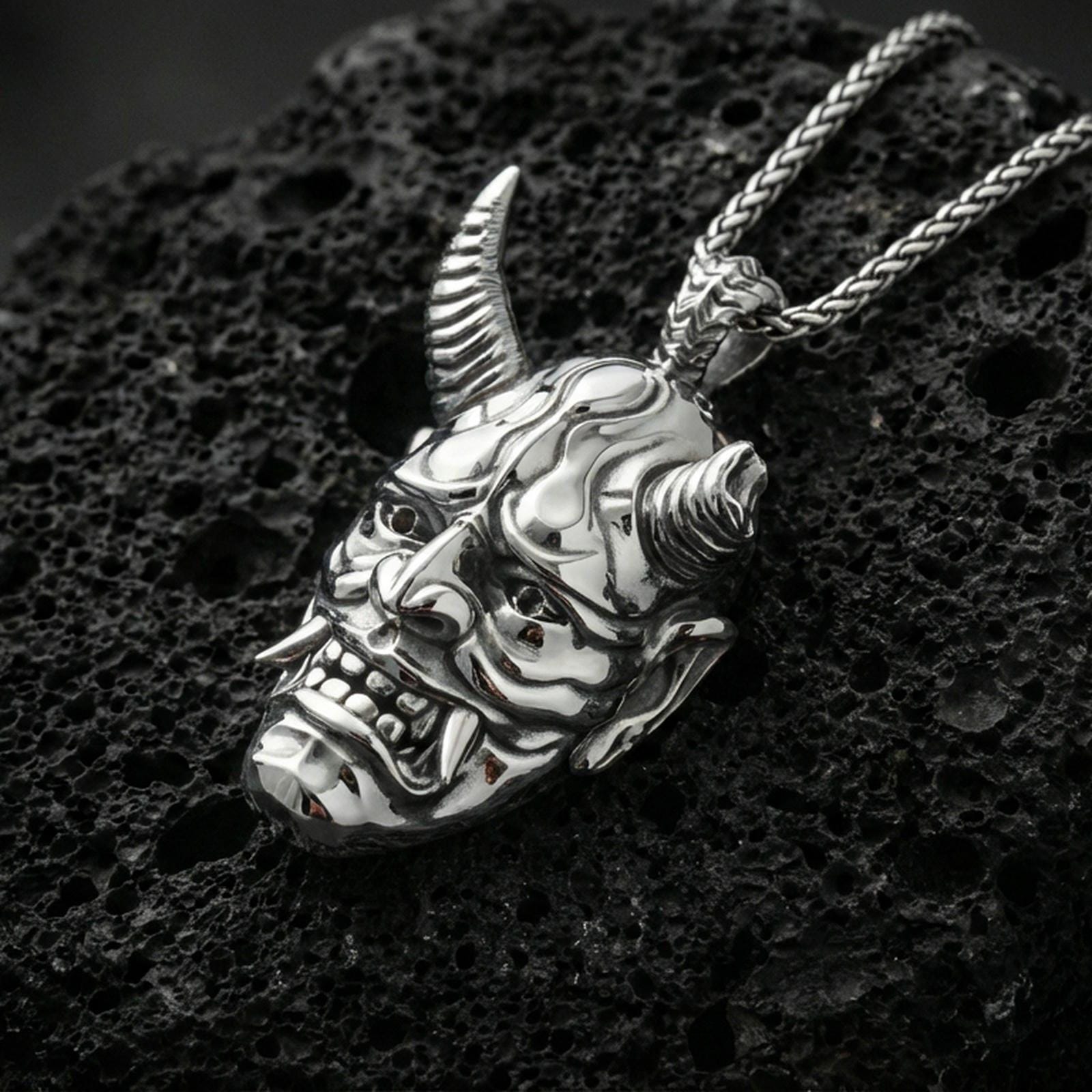 Sterling silver oni mask pendant in a polished silver finish, horned Japanese demon face with deep carved details, macro on volcanic stone