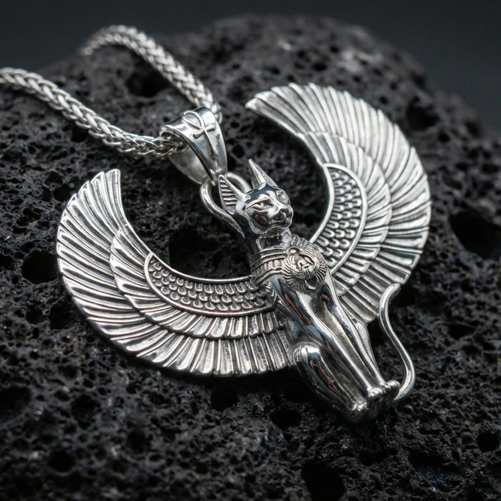 Sterling silver winged Bastet cat pendant in a polished silver finish, crisp feathered wings and sculpted cat face, macro on volcanic stone