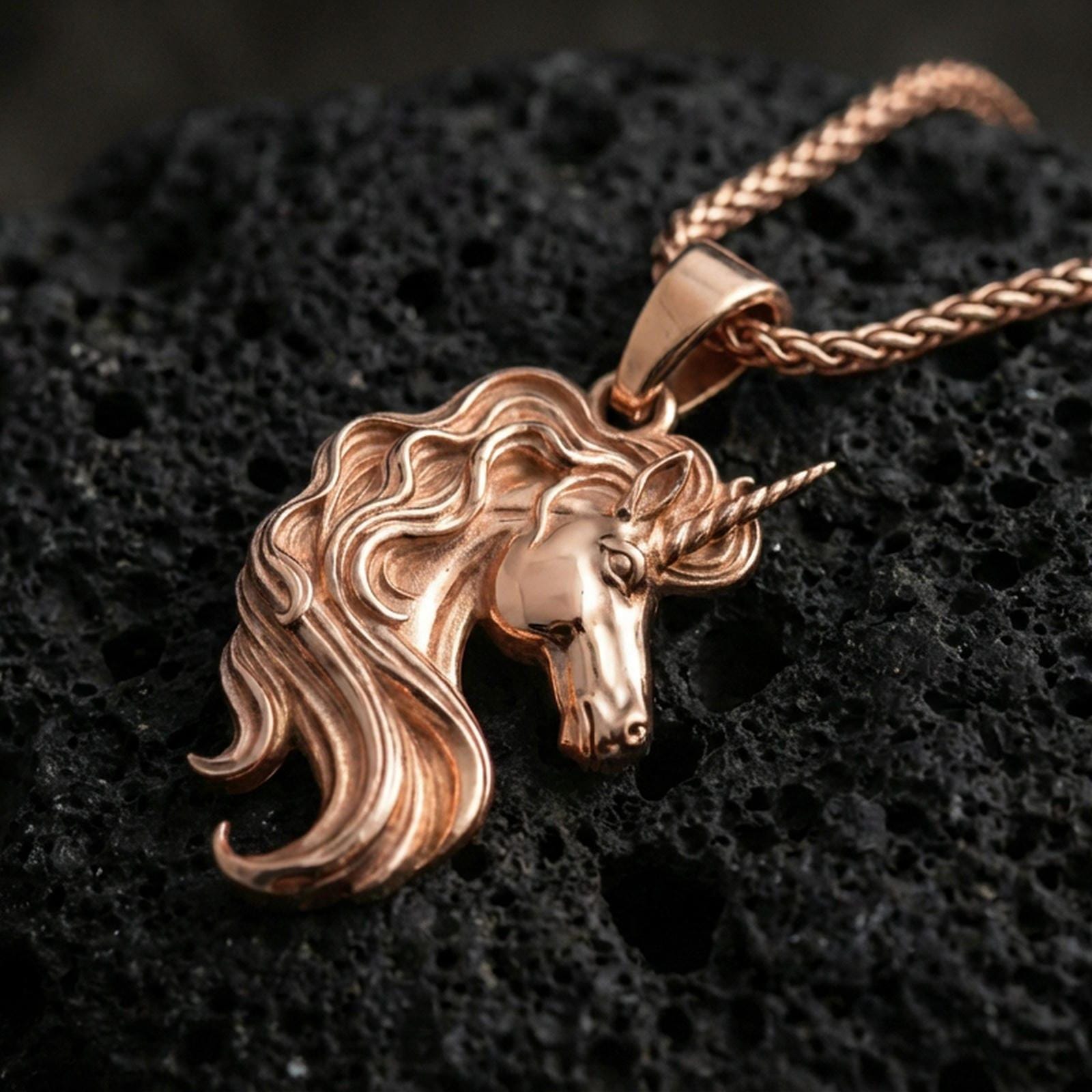 Rose gold plated unicorn head pendant necklace on textured volcanic rock, centered studio-style close-up showcasing a soft rosy glow on the unicorn profile and elegant mane lines.