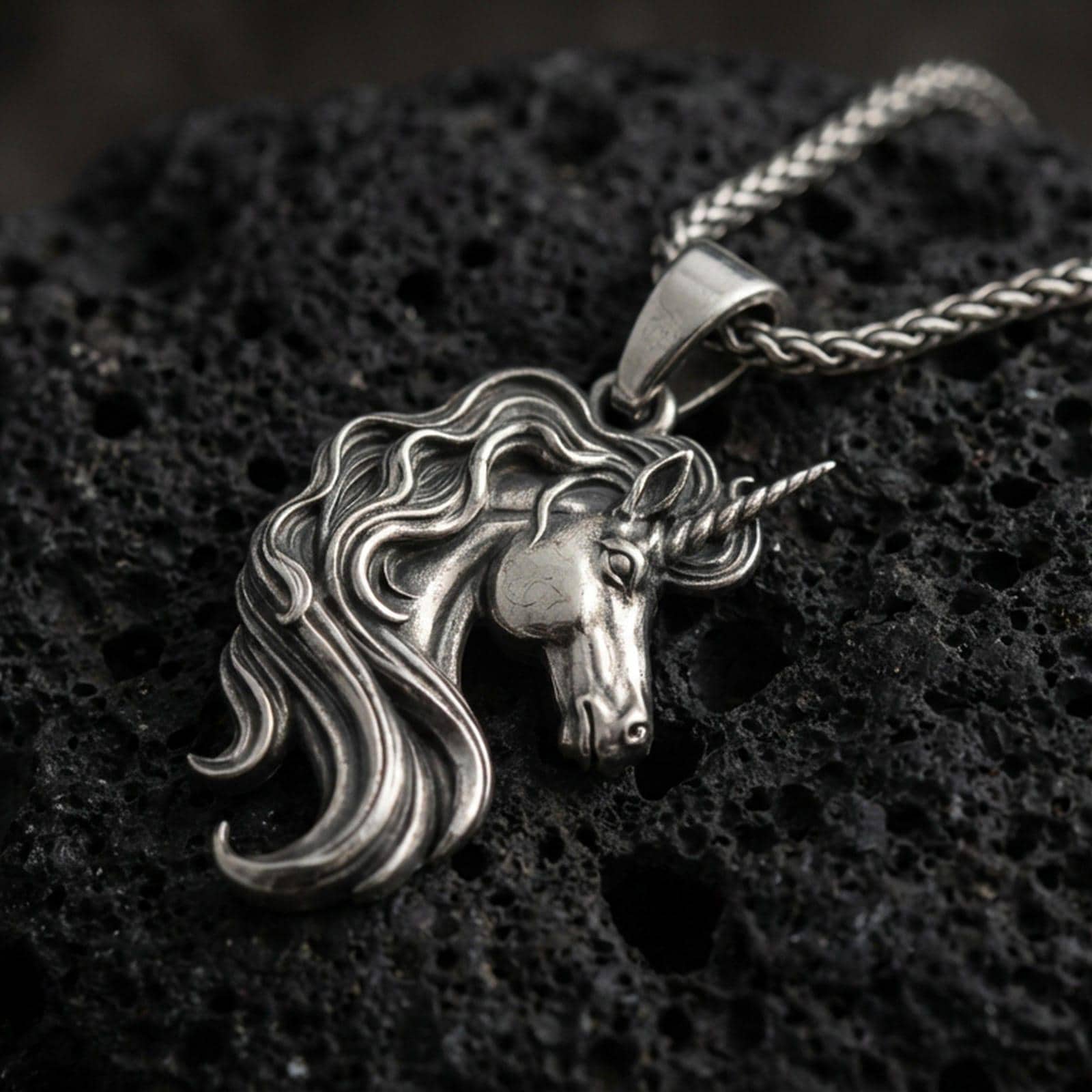 Oxidized unicorn head pendant necklace on textured volcanic rock, centered close-up highlighting the spiraled horn, flowing mane, and antique shadowed depth.