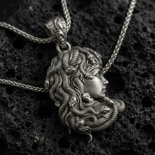 Oxidized Medusa profile pendant necklace on textured volcanic rock, centered close-up highlighting the layered snake hair, fine scale texture, and antique darkened depth.