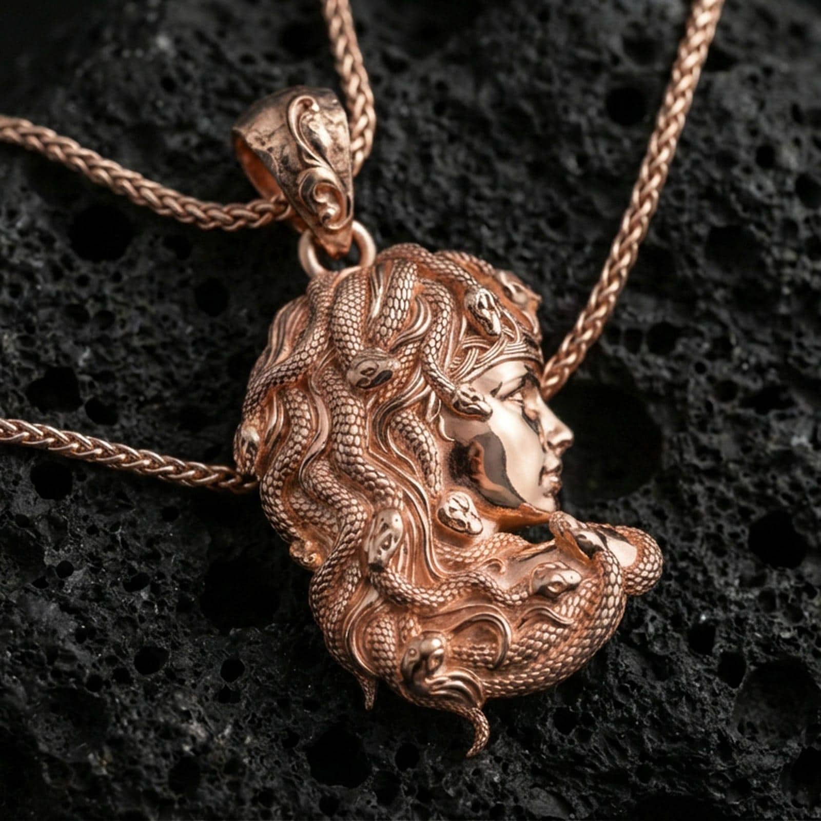 Rose gold plated Medusa profile pendant necklace on textured volcanic rock, centered studio-style close-up showcasing a soft rosy glow on the profile portrait and intertwined serpent detail.