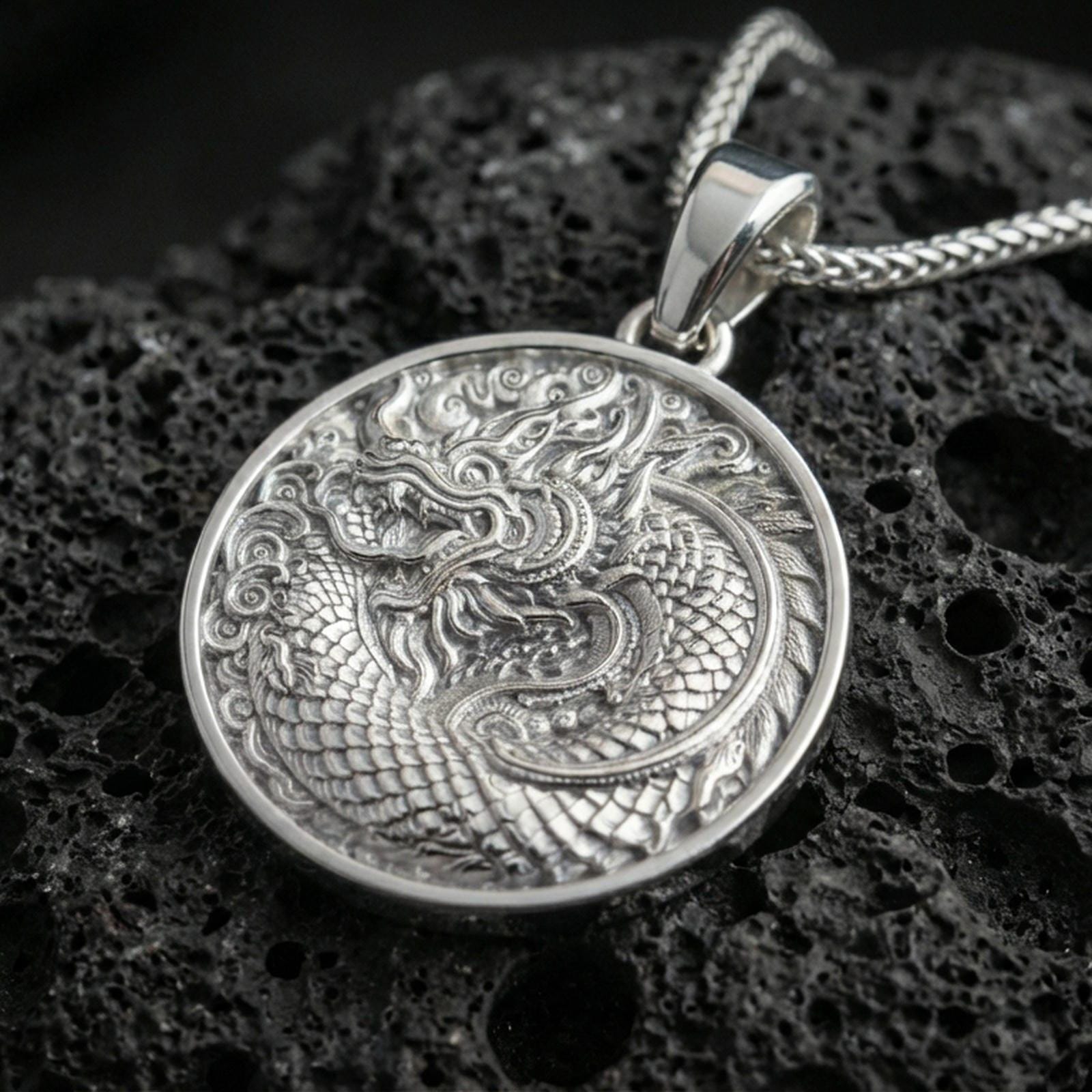 Polished silver oriental dragon medallion pendant necklace on textured volcanic rock, crisp macro product photo emphasizing sharp relief, clean edges, and detailed cloud swirls.