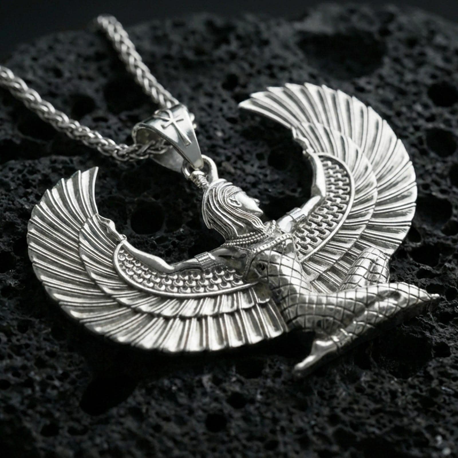 Polished silver winged goddess Isis pendant necklace on textured volcanic rock, crisp macro product photo emphasizing bright reflections, sharp feather detailing, and clean sculpted edges.